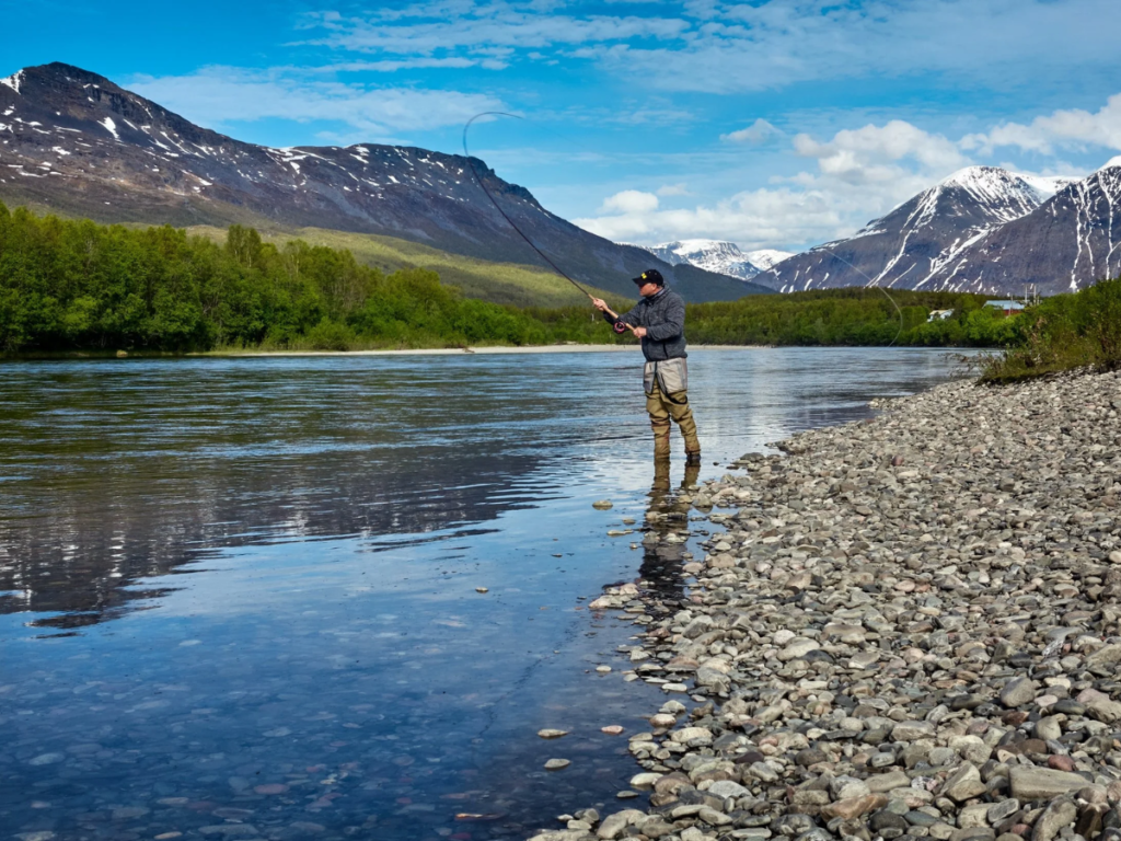 Fly fisherman casting in a mountain river near the Rocky Mountains with snowcapped peaks in the background.